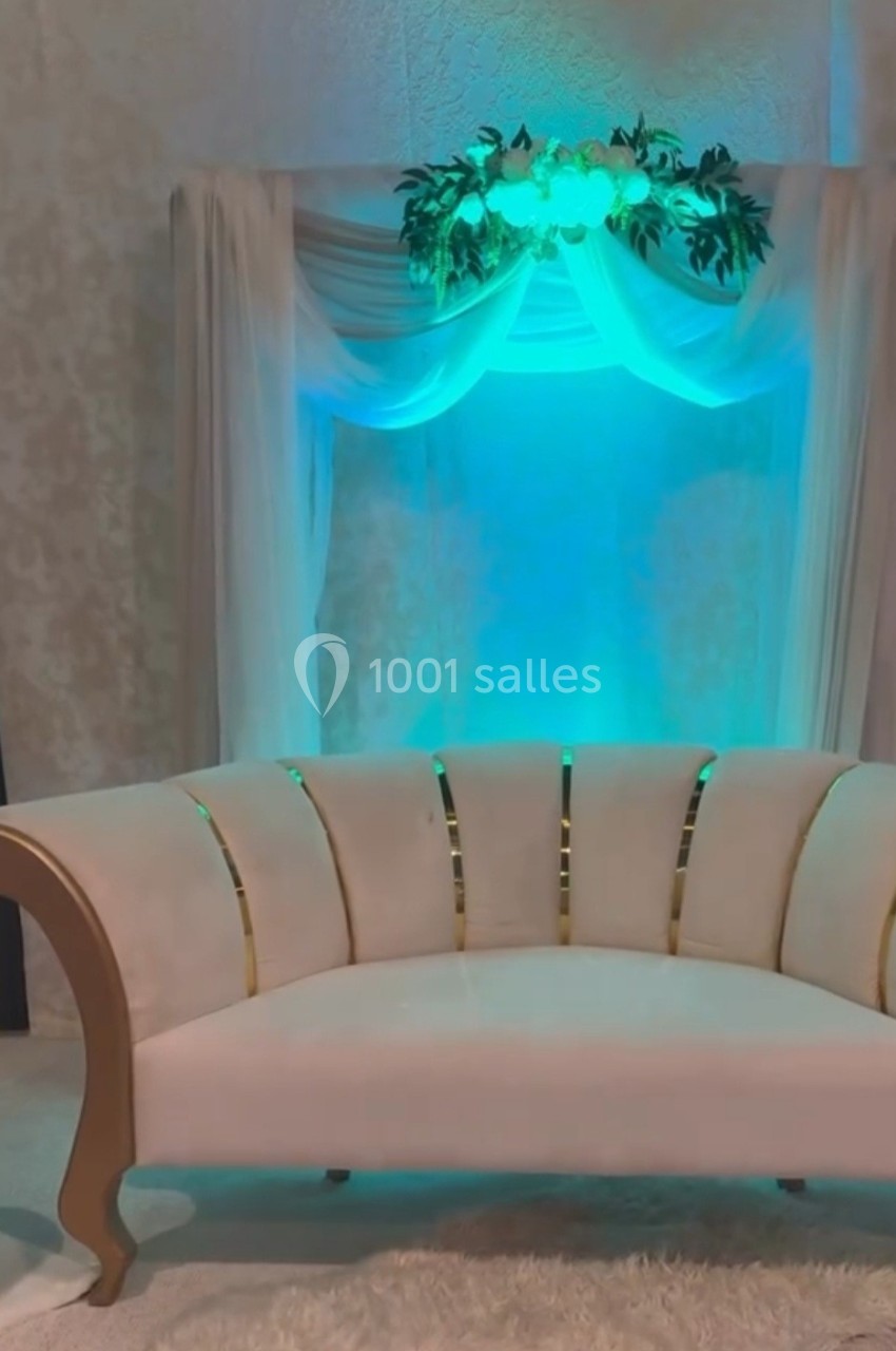 Canapé beige à accoudoirs courbés devant un rideau blanc drapé, éclairé par une lumière verte et décoré de feuillage.