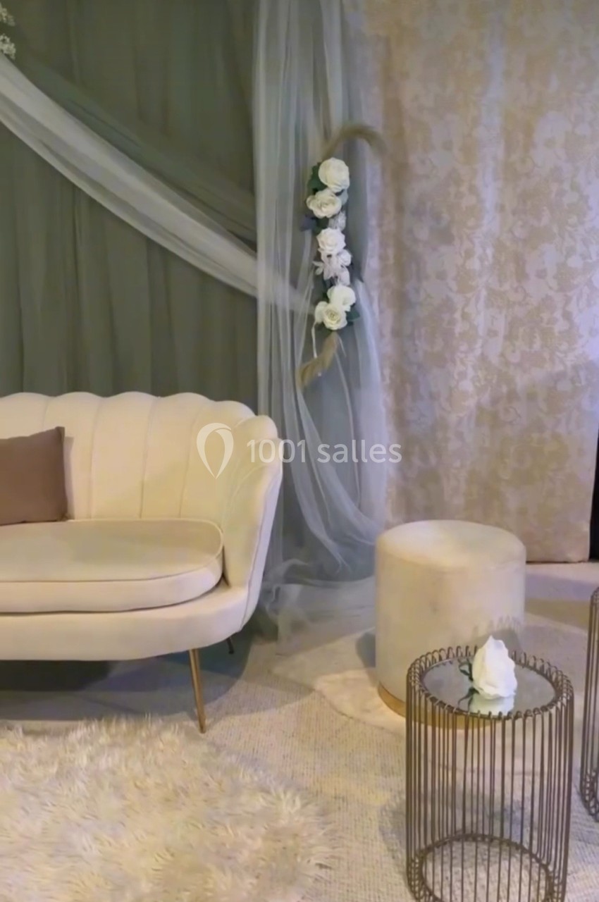 Salon élégant avec canapé beige, pouf assorti, table basse métallique et décor floral sur fond de rideaux.