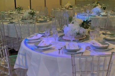 Salle de réception élégante avec tables rondes dressées, chaises transparentes et décorations florales sur chandeliers dorés.