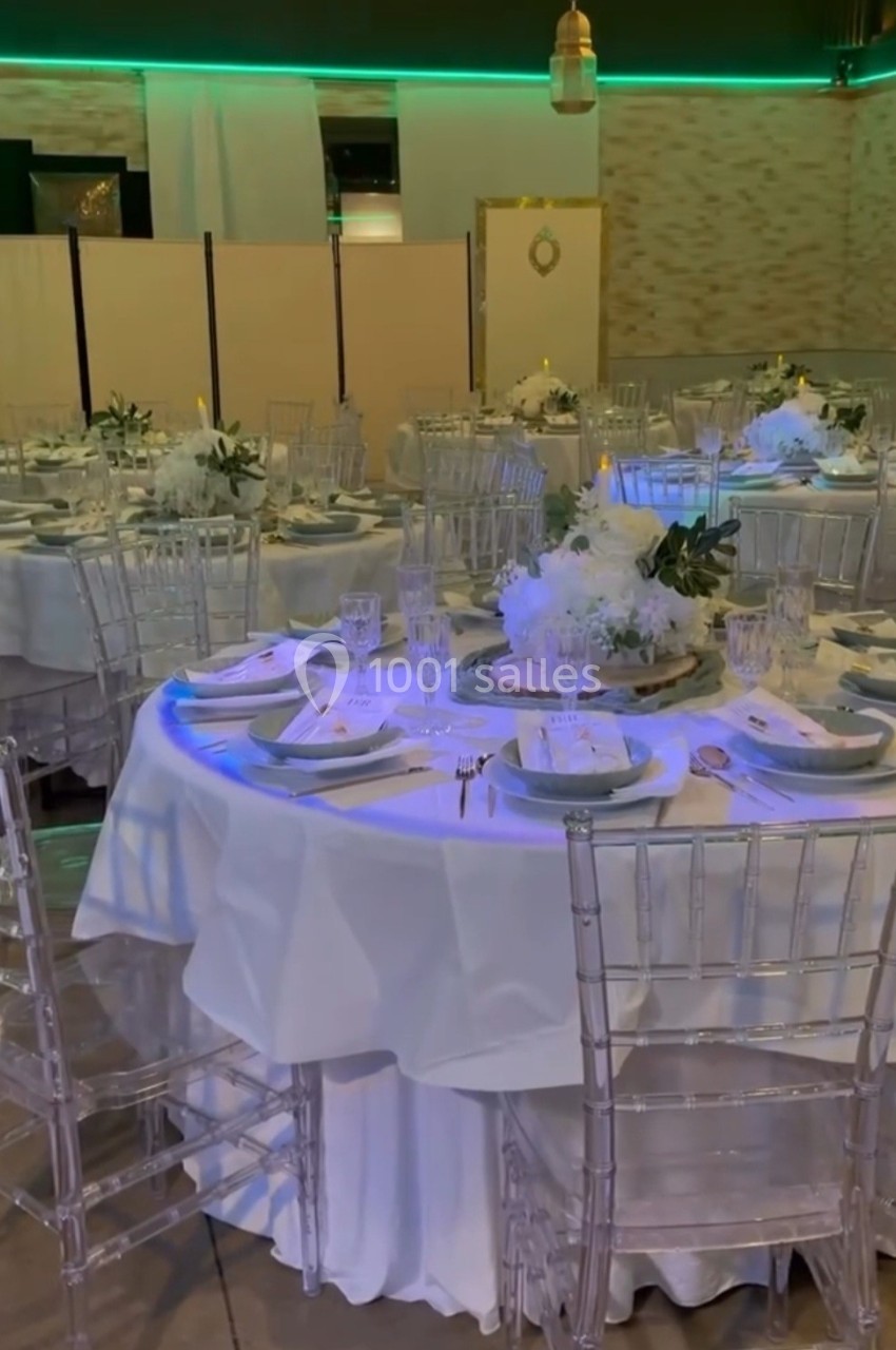 Salle de réception décorée avec des tables rondes, nappes blanches, chaises transparentes et centres de table fleuris.