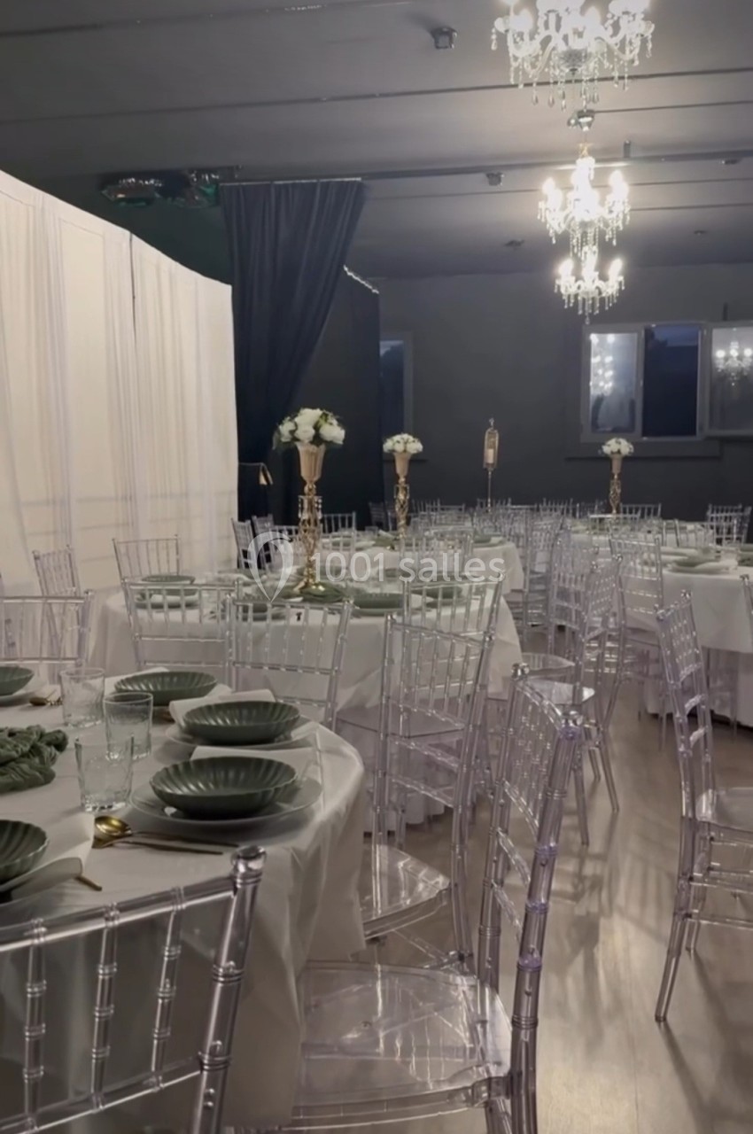 Salle de réception élégante avec tables rondes dressées, chaises transparentes et décorations florales sur chandeliers dorés.