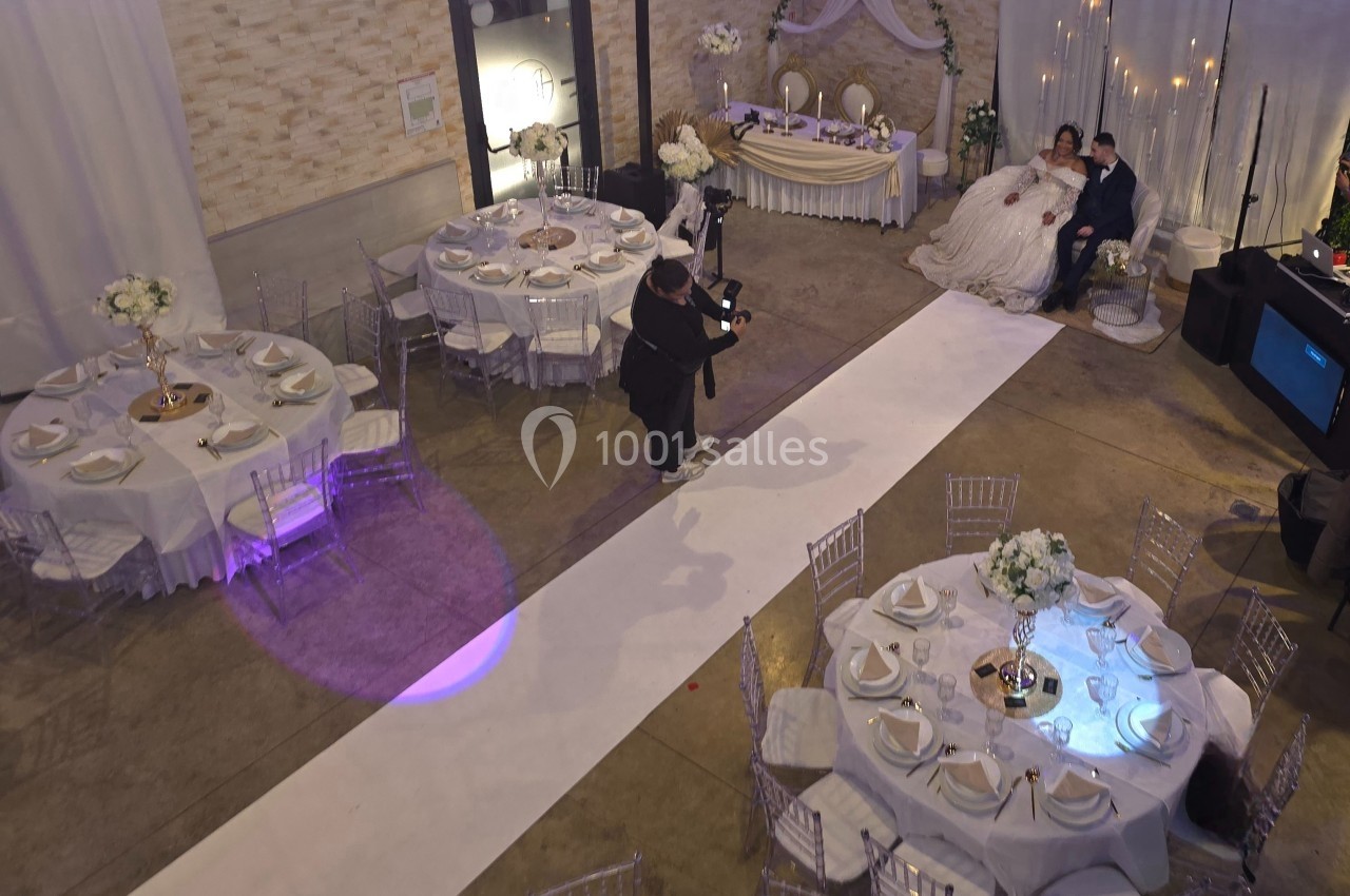 Salle de réception décorée pour un mariage, avec des tables dressées et un couple de mariés posant pour une photo.