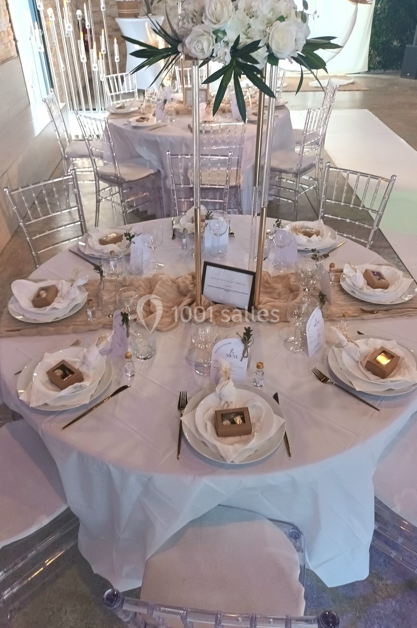 Table ronde élégamment dressée avec nappes blanches, chaises transparentes et décorations florales blanches au centre.