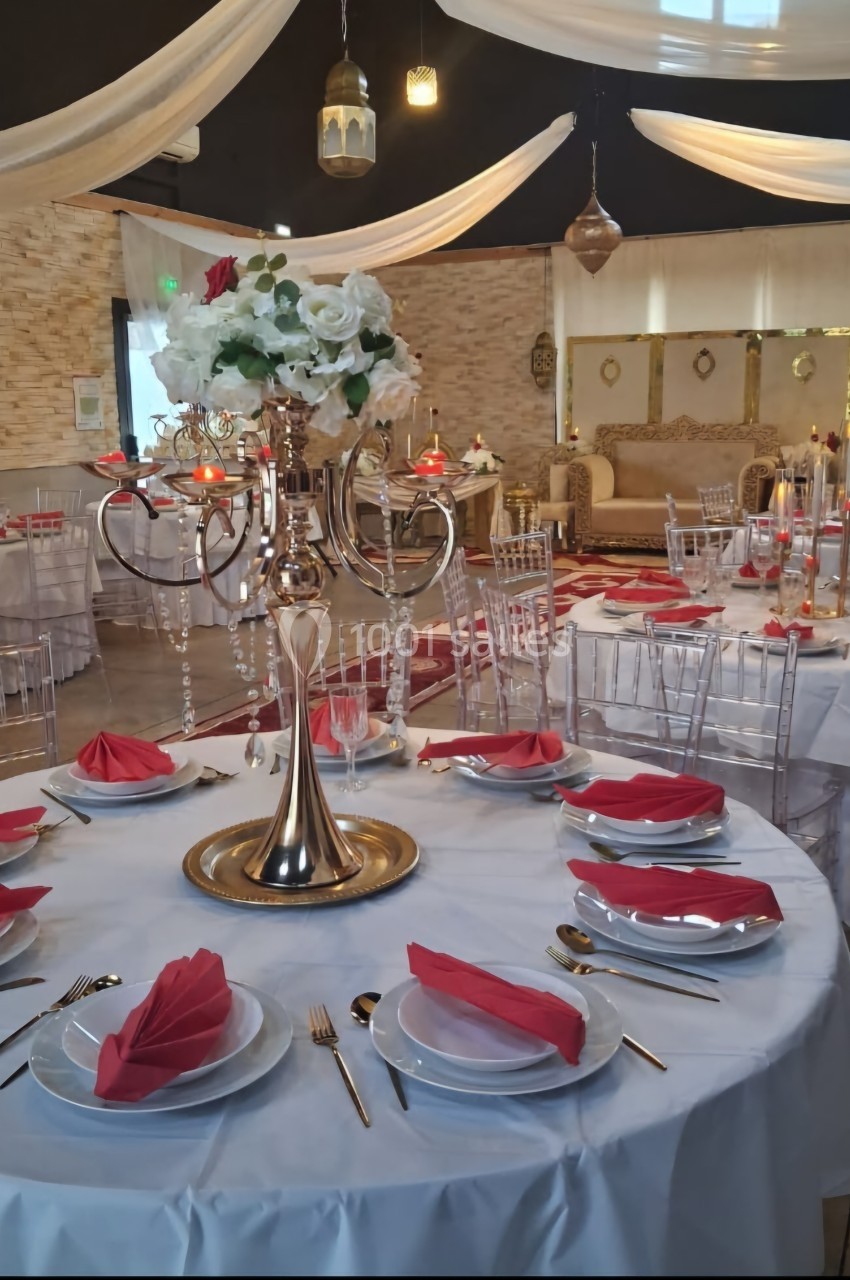 Salle de réception décorée avec des tables dressées, nappes blanches, serviettes rouges et un centre de table floral.