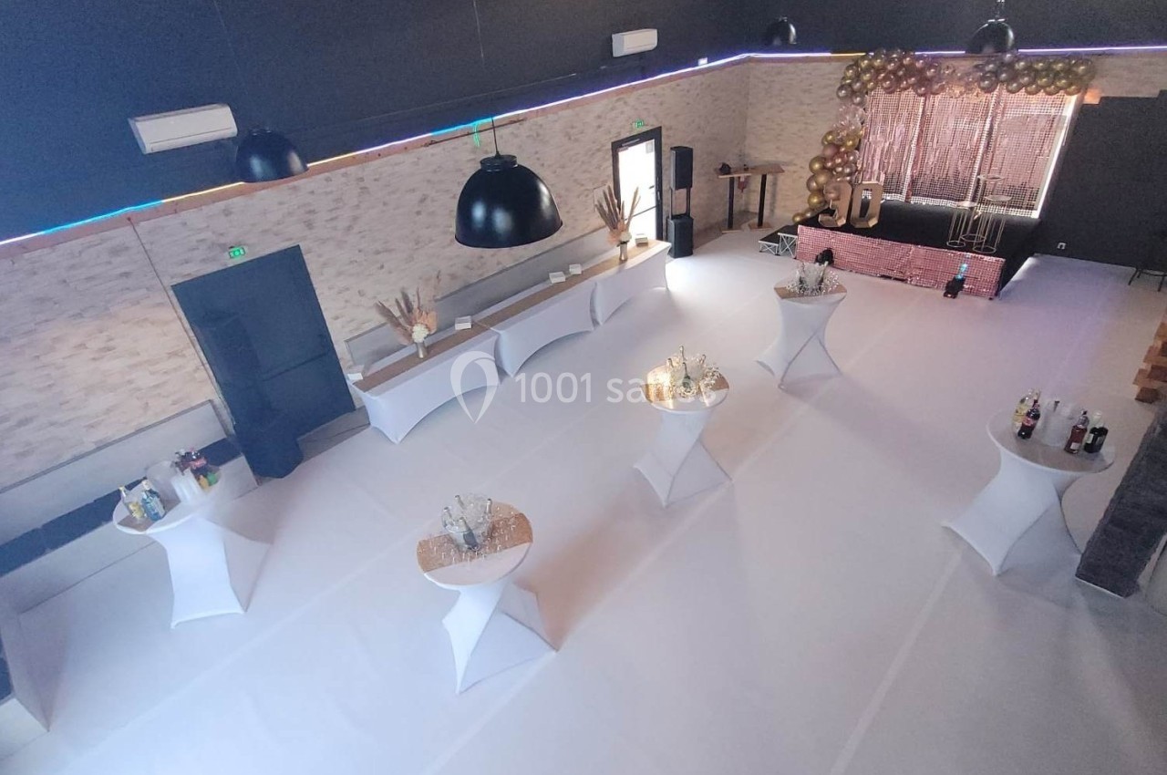Salle de réception décorée avec des tables hautes blanches, un espace scénique et des guirlandes lumineuses.