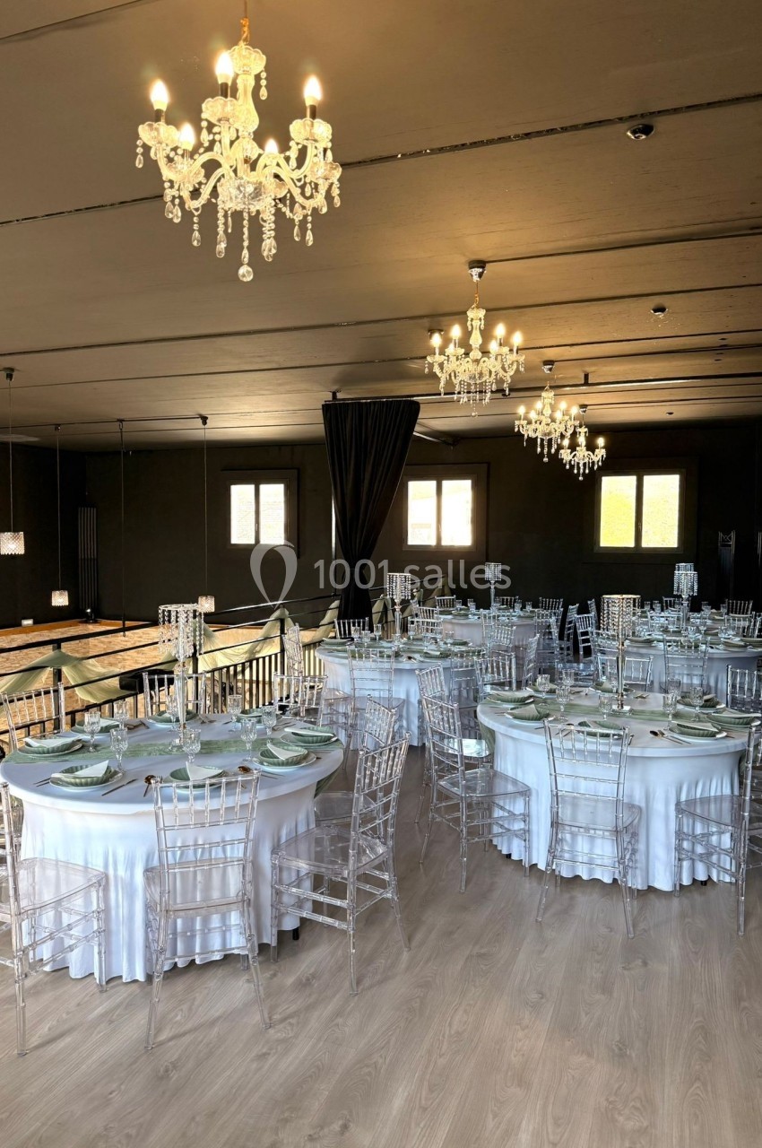 Salle de réception élégante avec tables rondes dressées, chaises transparentes et lustres suspendus.