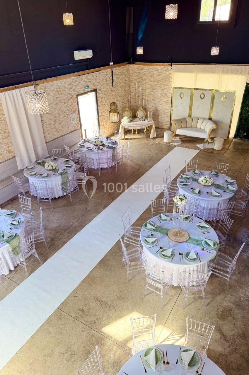 Salle de réception décorée avec des tables rondes dressées, des chaises transparentes et une allée centrale blanche.