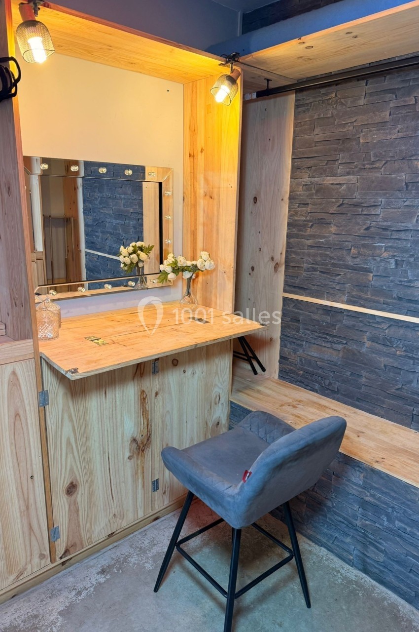 Coiffeuse en bois avec miroir éclairé, chaise grise et mur en pierre décorative en arrière-plan.