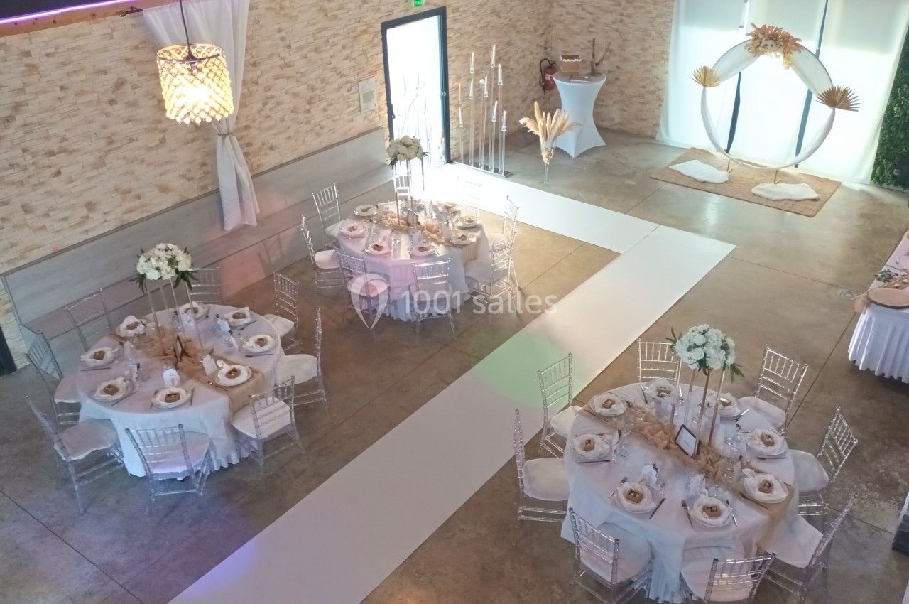 Salle de réception décorée avec des tables rondes, nappes blanches, chaises transparentes et une arche florale en arrière…
