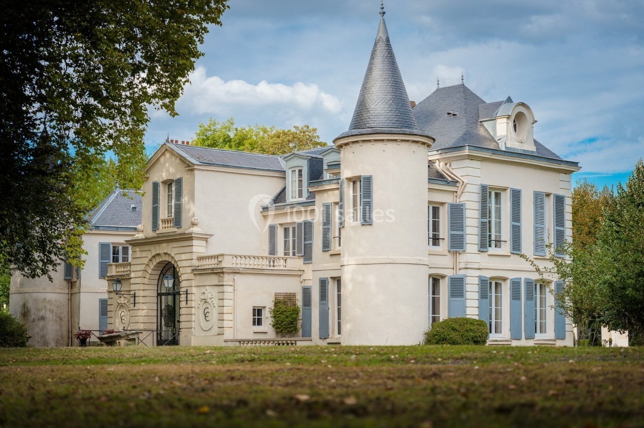 Grande maison blanche avec tour ronde, volets bleus et jardin, entourée d'arbres sous un ciel partiellement nuageux.
