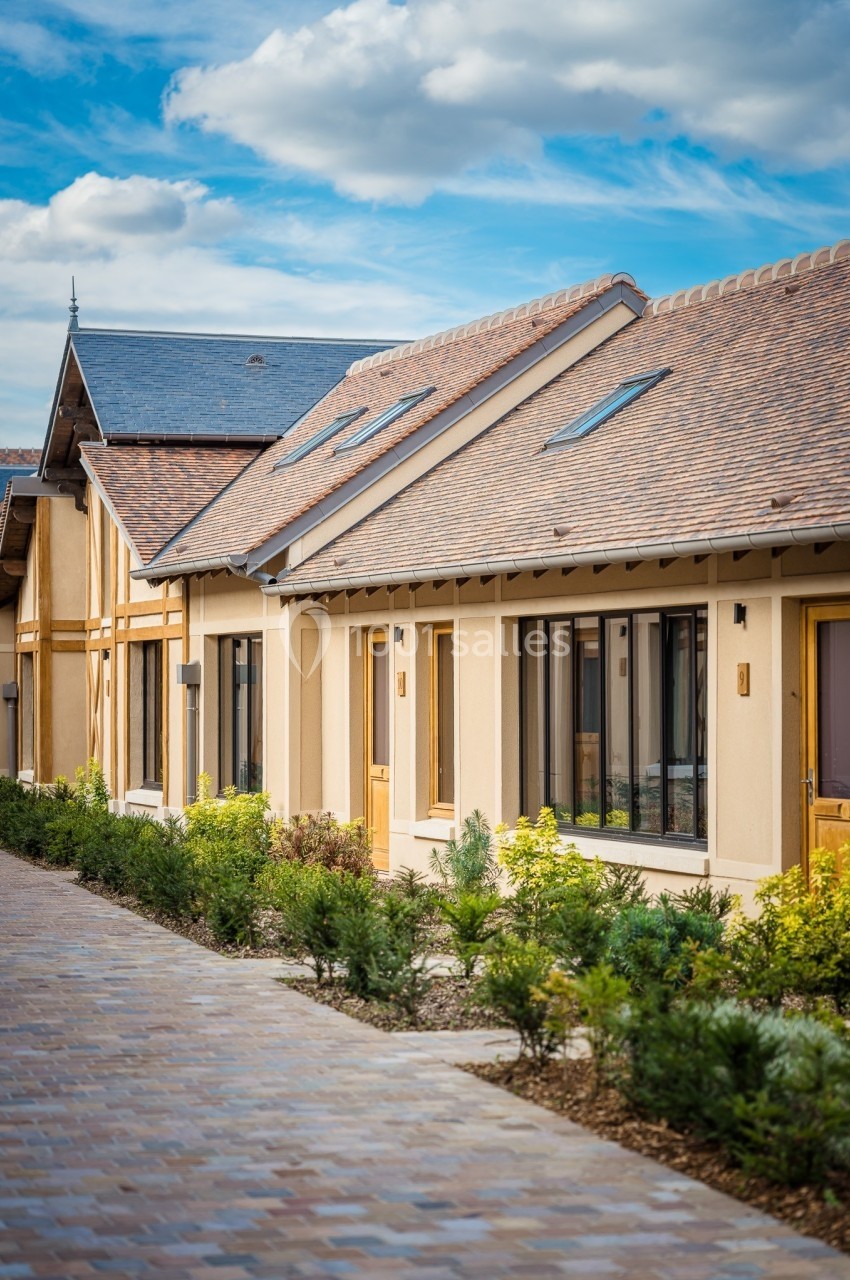 Façade d'un bâtiment moderne avec toit en tuiles, grandes fenêtres et allée pavée bordée de végétation.