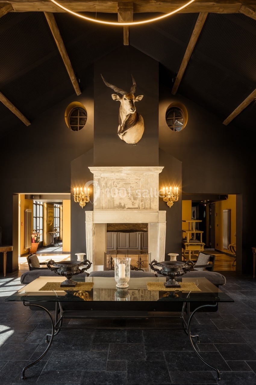 Salle de séjour sombre avec cheminée en pierre, tête de cerf au mur, table en verre et poutres apparentes au plafond.