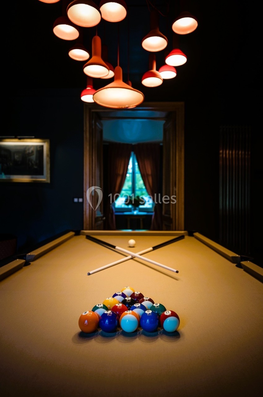 Table de billard éclairée avec des boules colorées alignées et deux queues croisées, dans une pièce sombre et élégante.