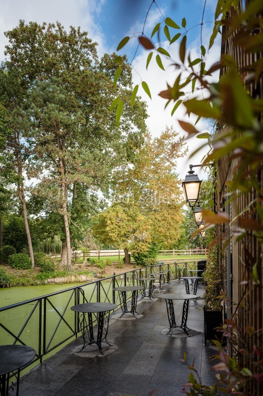 Terrasse avec tables et chaises en métal noir, bordée de végétation, donnant sur un étang entouré d'arbres.