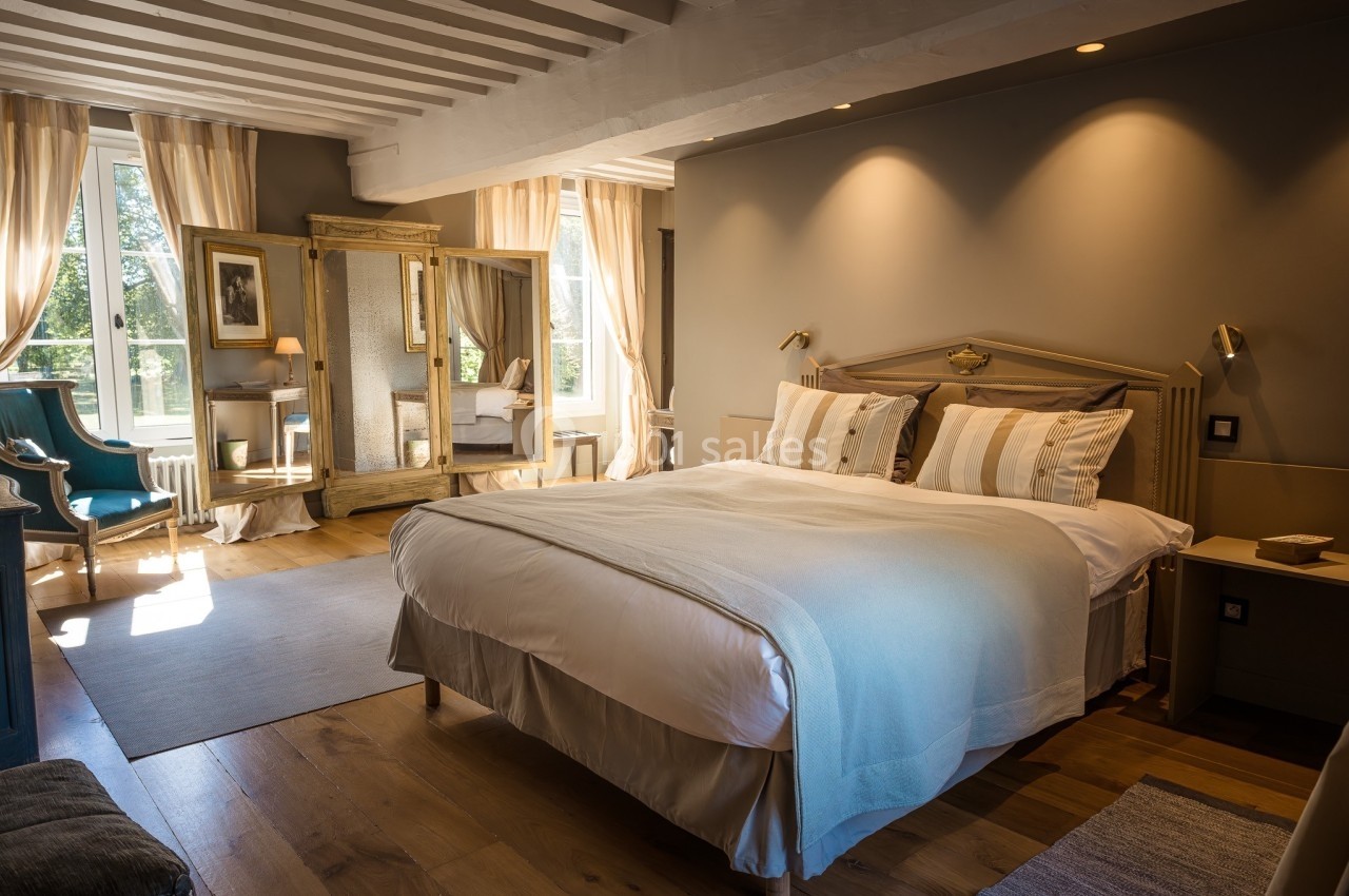 Chambre lumineuse avec lit double, parquet en bois, fauteuil, miroir ancien et grandes fenêtres avec rideaux beiges.