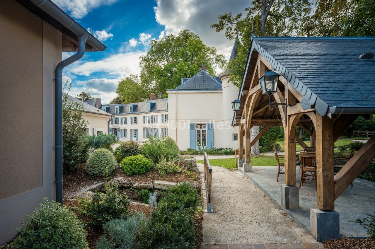 Cour d'un domaine avec un bâtiment principal aux façades claires, jardin paysager et pergola en bois sous un ciel…
