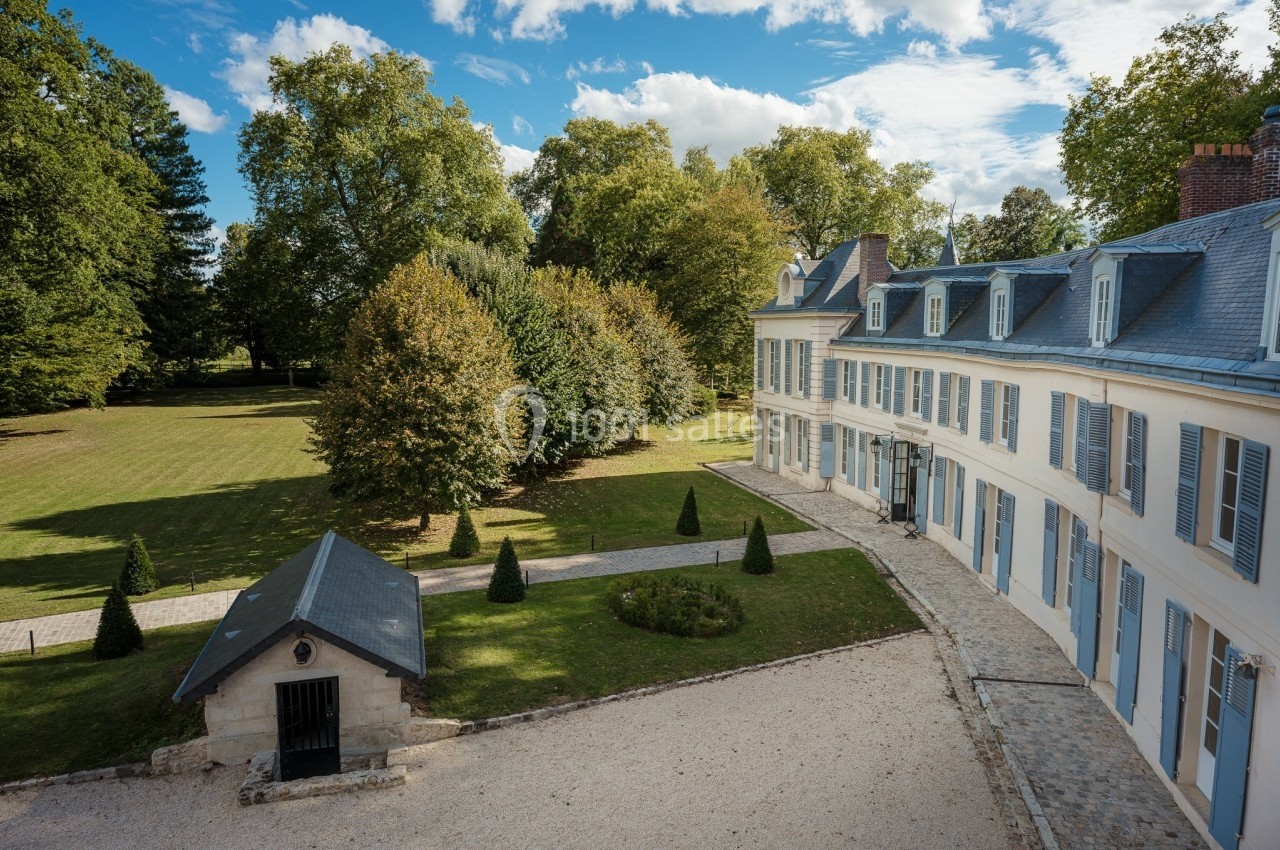 Vue d'un bâtiment historique entouré d'un parc arboré avec une allée pavée et une petite dépendance au premier plan.