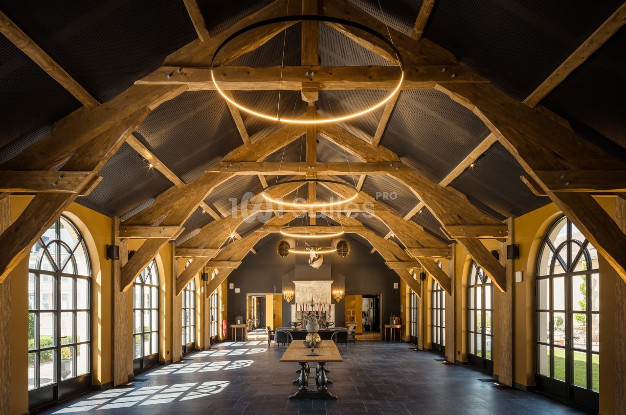 Salle spacieuse avec charpente en bois apparente, grandes fenêtres cintrées et table centrale éclairée par des luminaires…