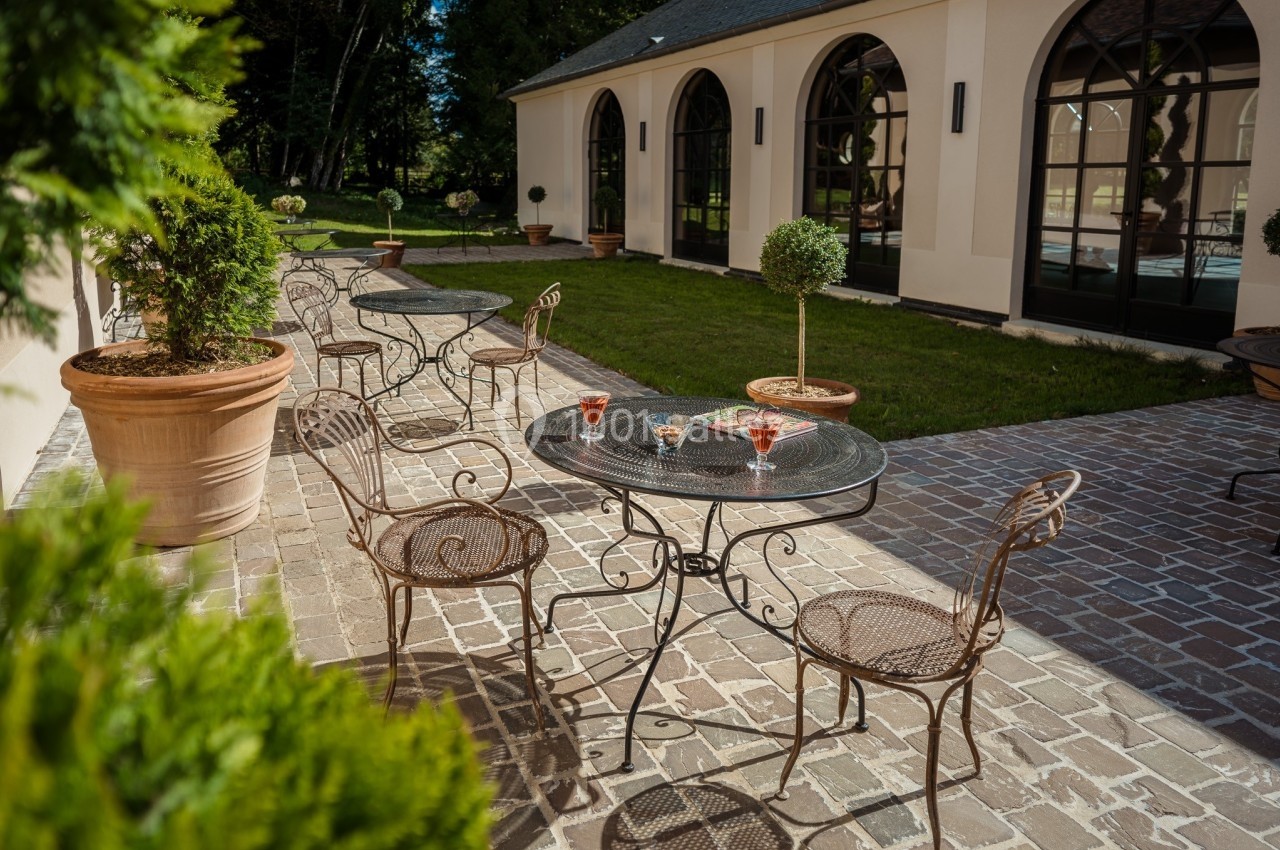 Terrasse pavée avec tables et chaises en fer forgé, verres posés, entourée de verdure et d'un bâtiment aux grandes fenêtres.