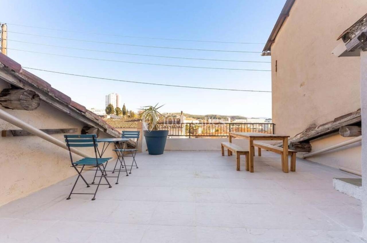 Terrasse en hauteur avec table, banc, chaises pliantes et plantes en pot, offrant une vue dégagée sur des toits et le ciel.