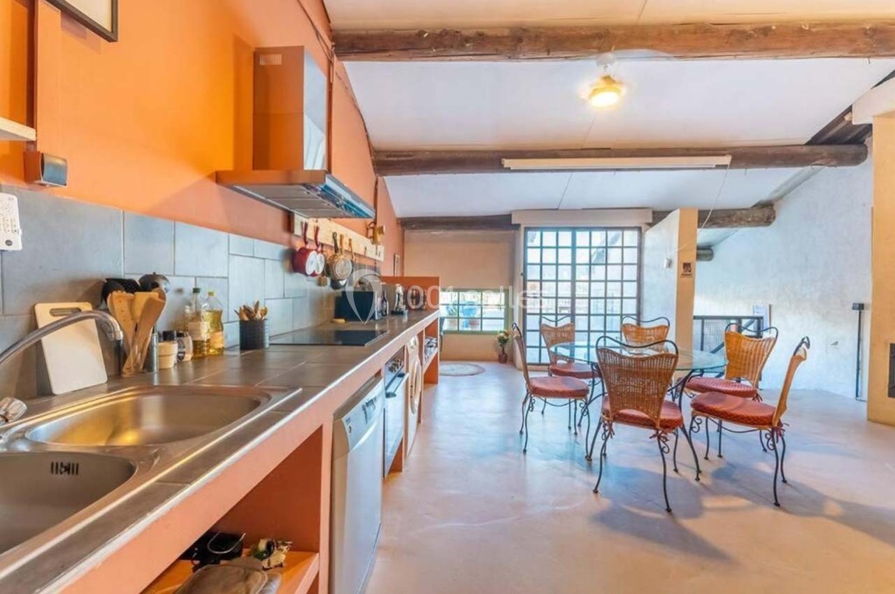 Cuisine spacieuse avec plan de travail en inox, murs orange, table avec chaises en osier et poutres apparentes.