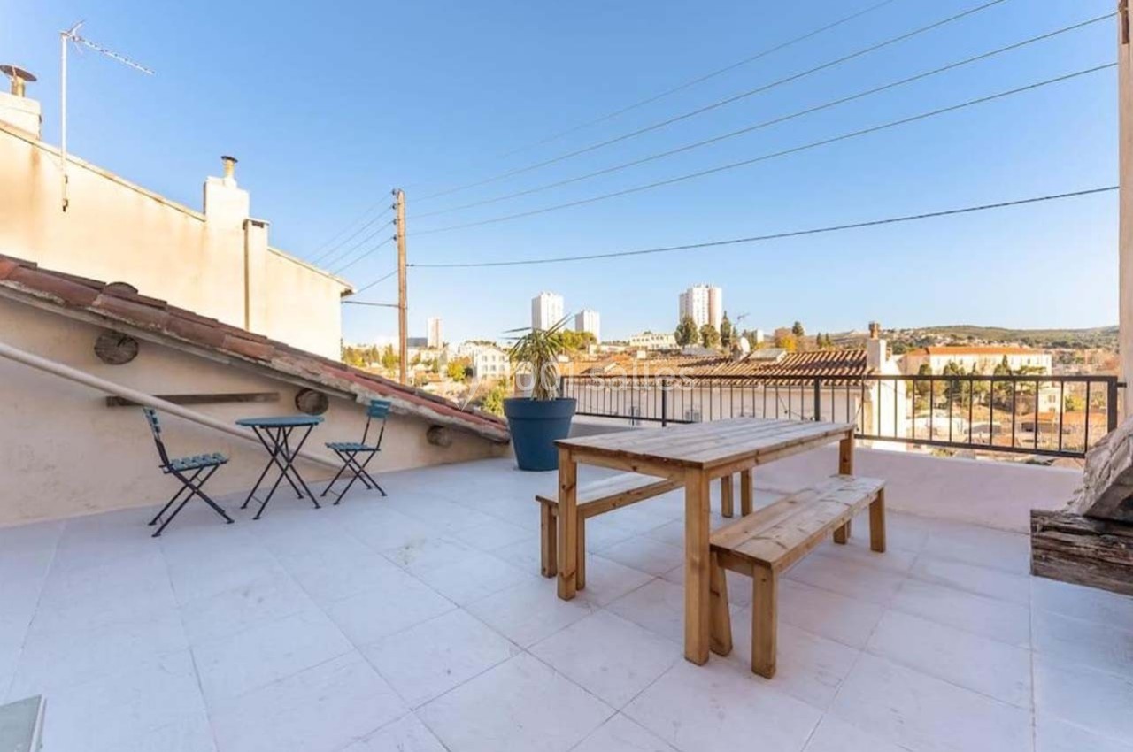 Terrasse ensoleillée avec table en bois, chaises pliantes et vue sur des bâtiments et collines au loin.
