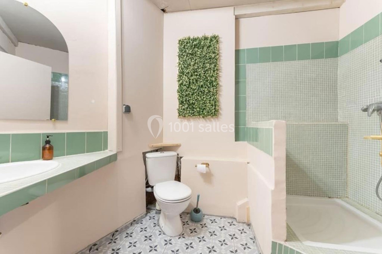 Salle de bain avec toilettes, lavabo, douche à l'italienne, carrelage vert et décoration murale végétale.
