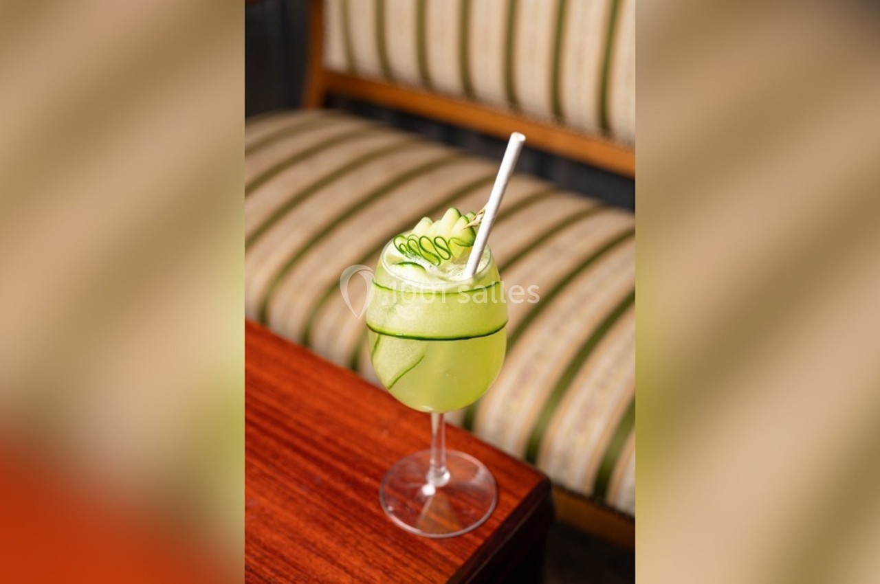 Verre de cocktail vert garni de tranches de concombre, posé sur une table en bois devant une banquette rayée.