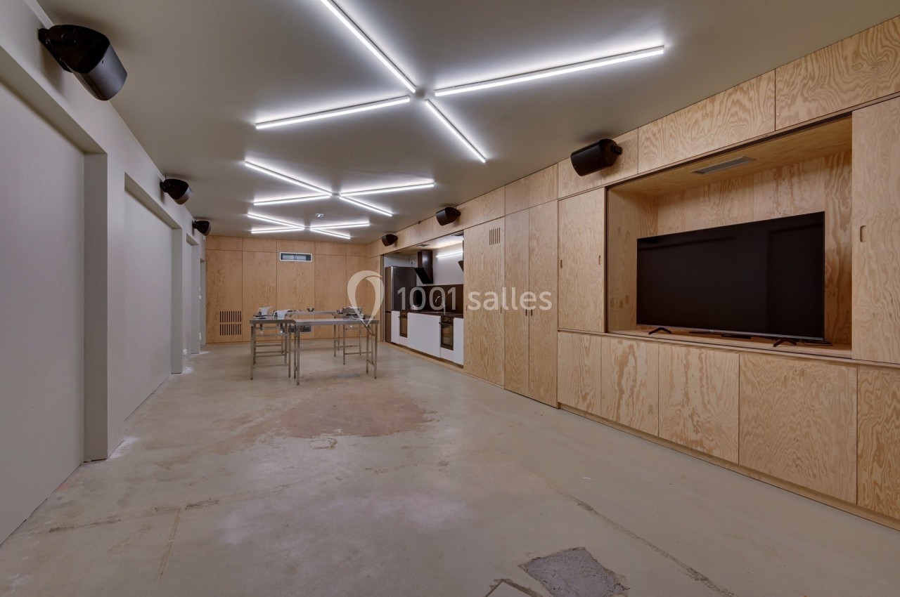 Salle moderne avec murs en bois clair, éclairage géométrique au plafond, tables en métal et écran encastré.