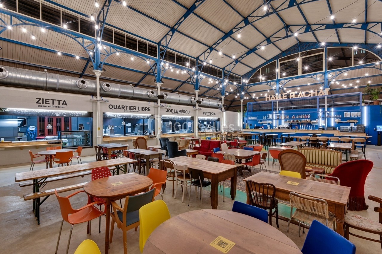 Intérieur d'une halle avec des tables en bois, des chaises colorées et des stands de restauration éclairés.