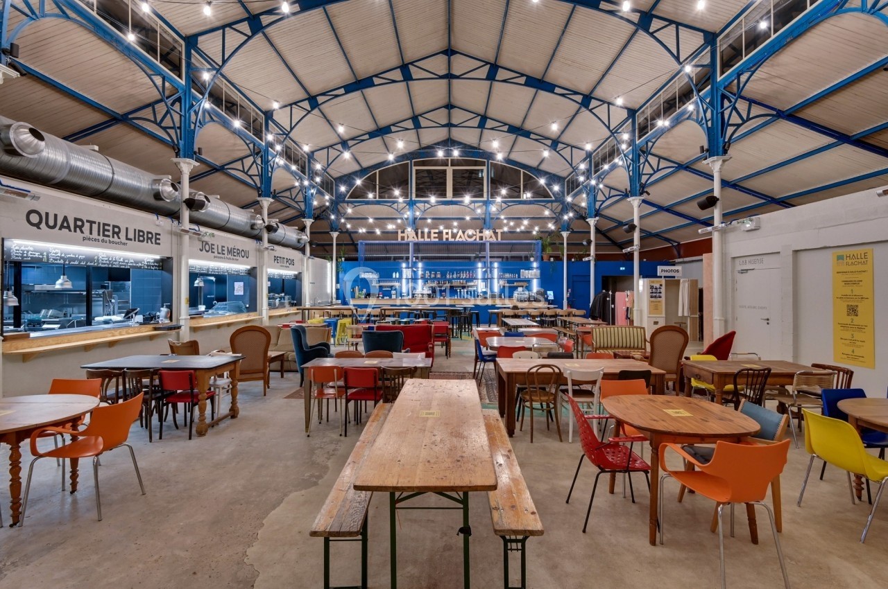 Vue intérieure d'une halle avec des tables et chaises colorées, un plafond voûté et des stands de restauration.
