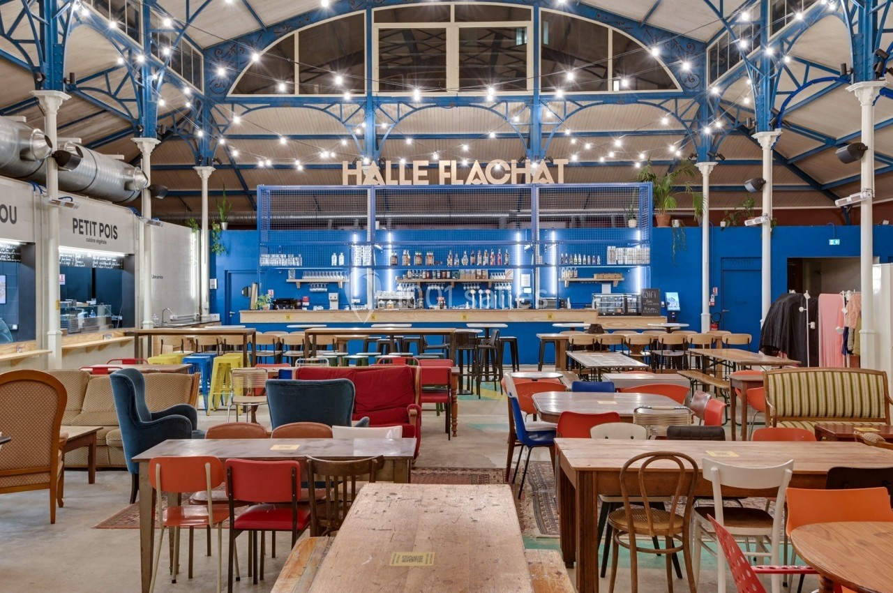 Intérieur d'une halle avec un bar central, des tables et des chaises variées dans un espace lumineux et coloré.