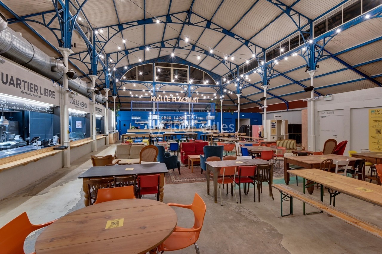 Intérieur d'une halle avec tables, chaises variées et stands de restauration sous une structure métallique éclairée.