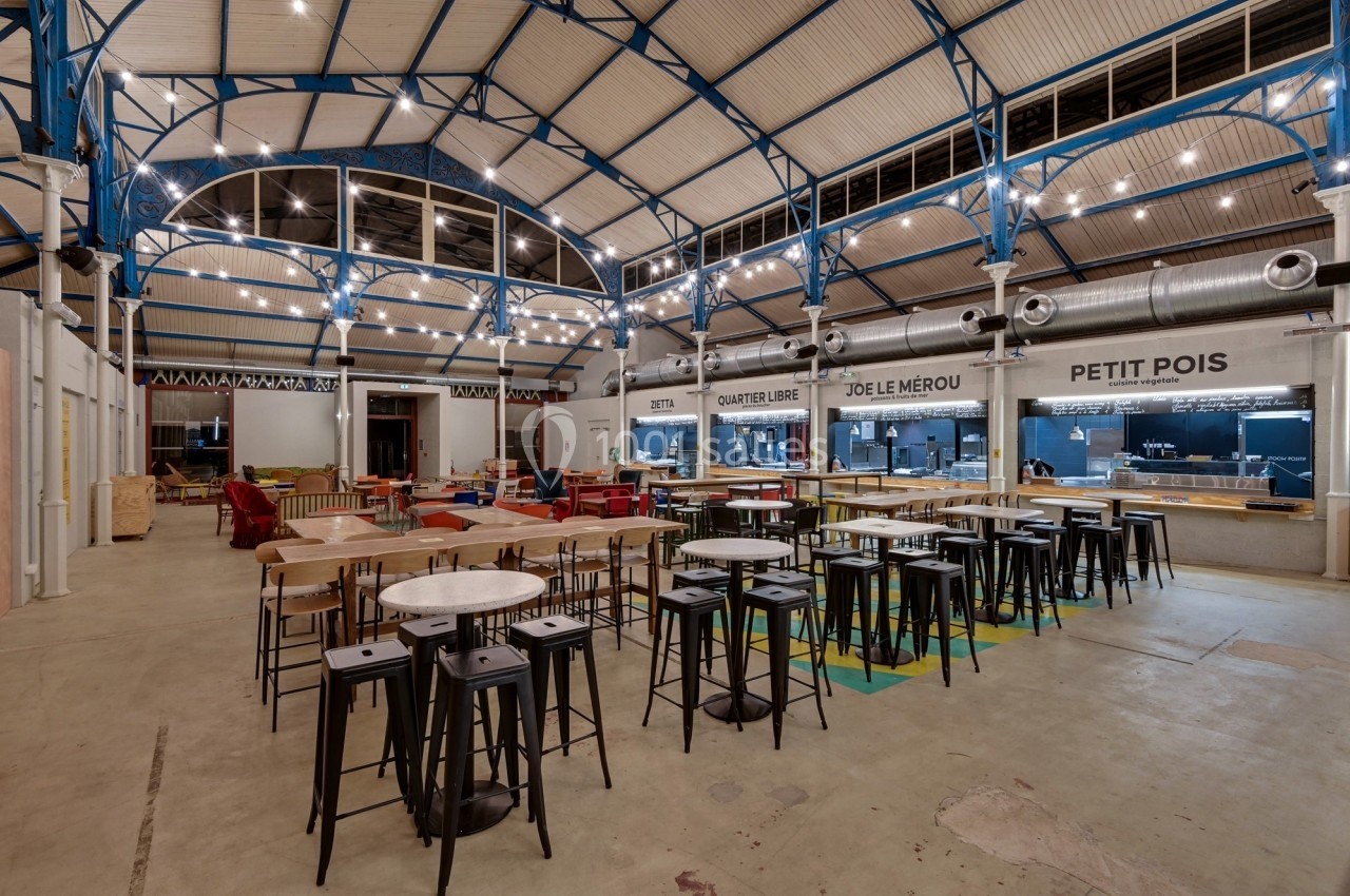 Intérieur d'un marché couvert avec tables, chaises et stands de restauration sous une structure métallique éclairée.