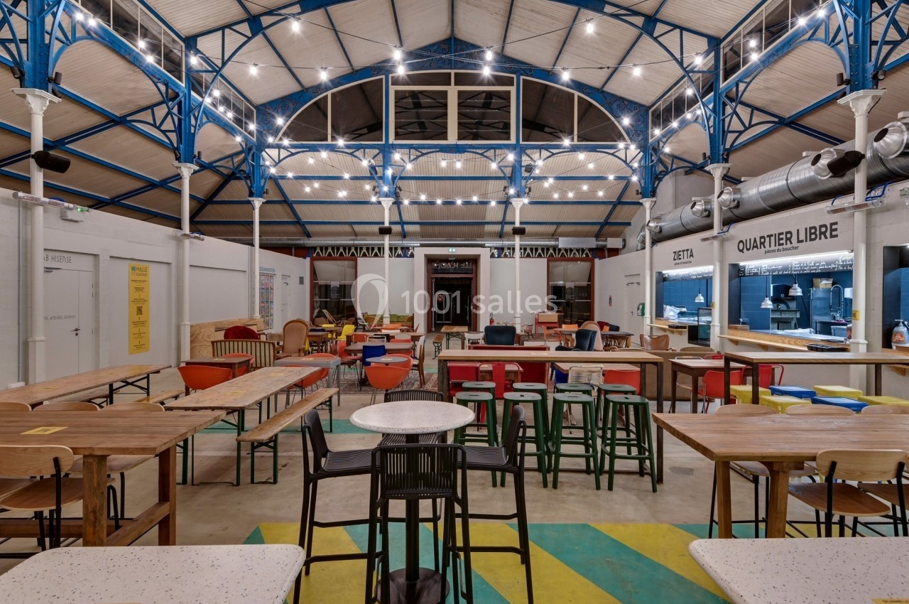 Intérieur d'une halle avec tables en bois, chaises colorées et stands de restauration sous une structure métallique éclairée.