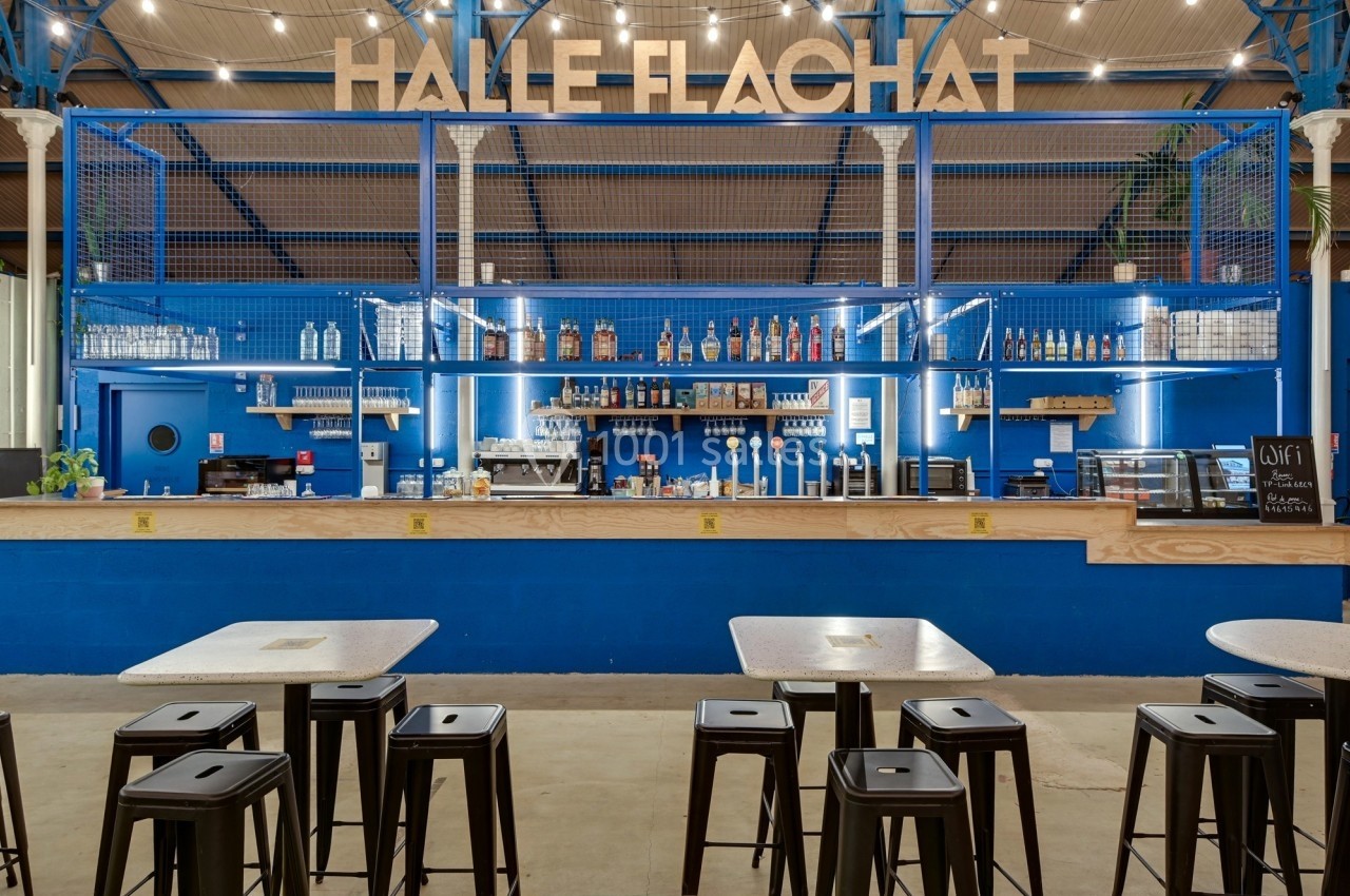 Bar moderne avec comptoir en bois, étagères illuminées, tabourets noirs et tables dans un espace industriel lumineux.