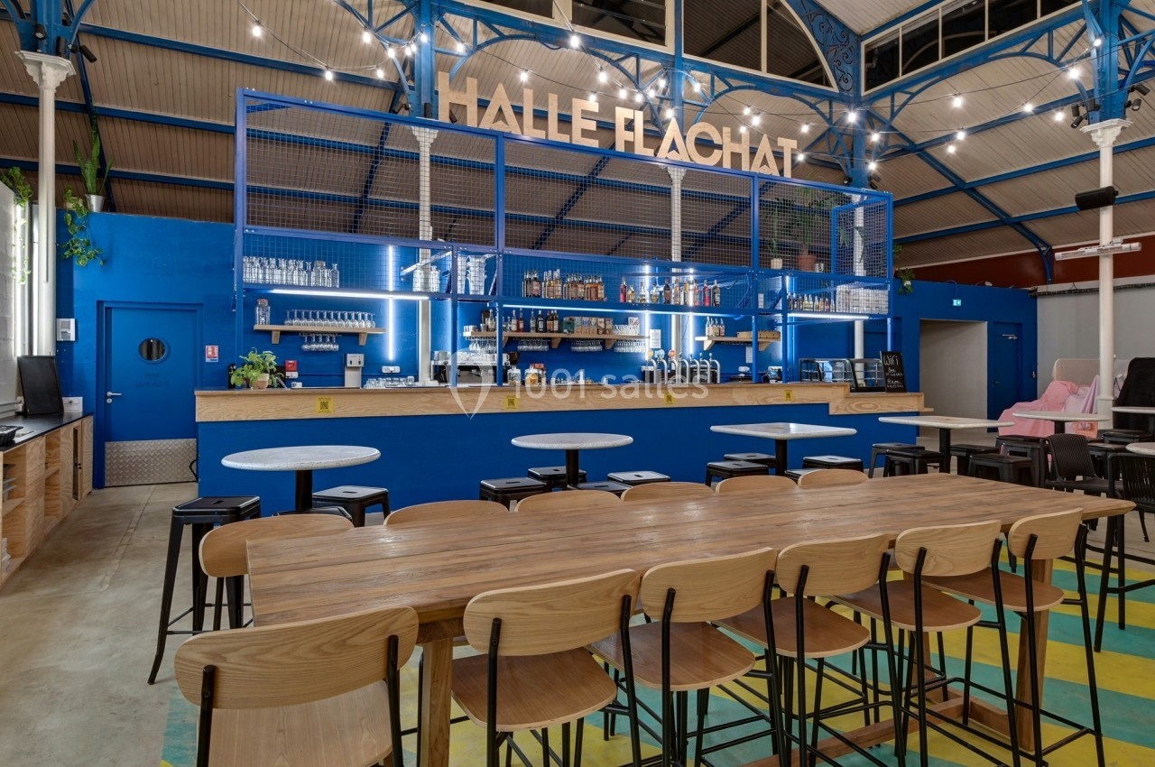 Intérieur d'une halle rénovée avec un bar moderne bleu, tables en bois et chaises alignées sous un plafond métallique.