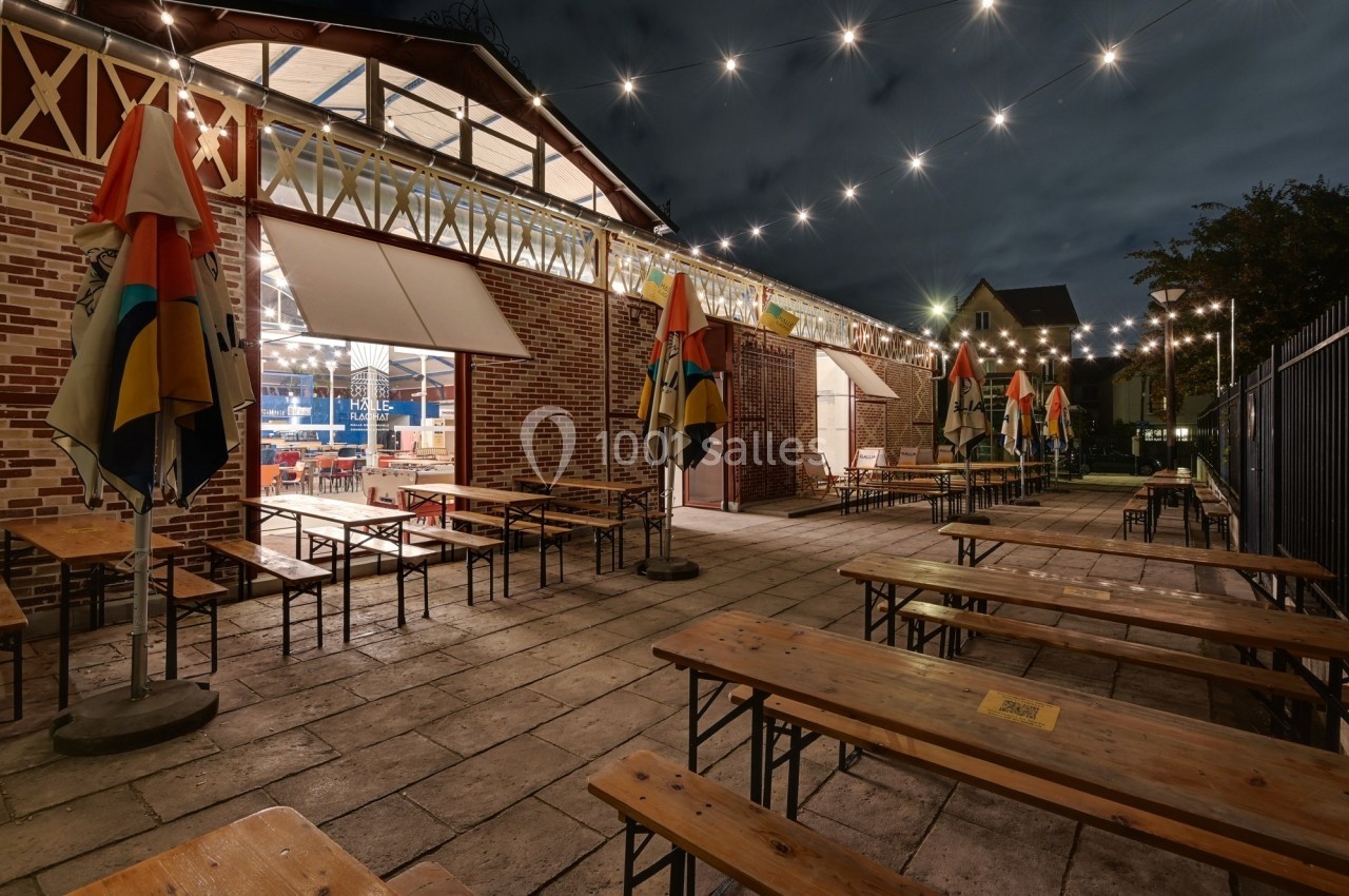 Terrasse extérieure avec tables en bois, éclairée par des guirlandes lumineuses, devant un bâtiment en briques.