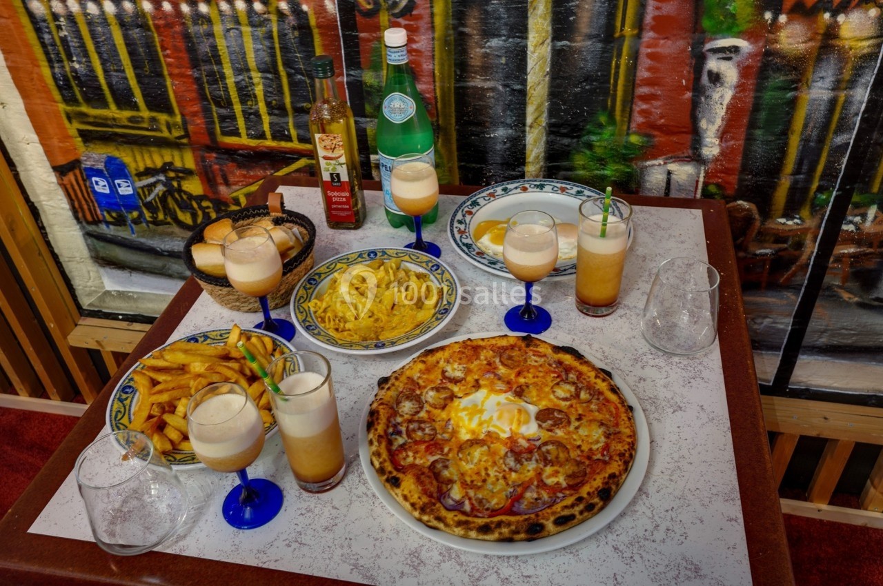 Table dressée avec une pizza, des frites, des boissons servies dans des verres et des bouteilles, devant une fresque colorée.