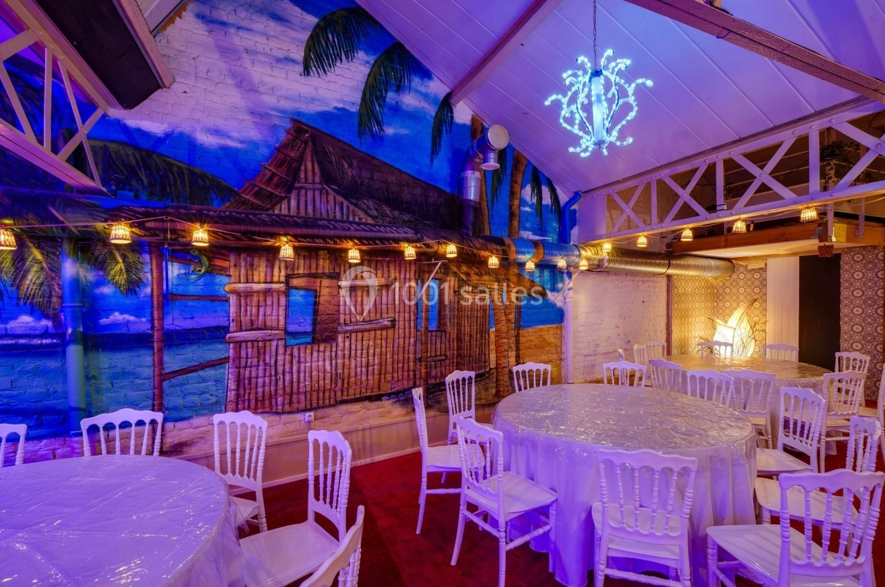 Salle décorée avec des tables rondes, chaises blanches et fresque murale tropicale sous un plafond avec poutres apparentes.
