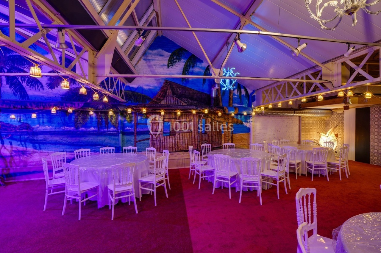 Salle décorée avec des tables rondes et chaises blanches, éclairage chaleureux et fresque murale tropicale.