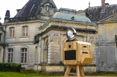 Miniature M-Photobooth #2 Borne photo en bois sur trépied, équipée d'un écran et d'un objectif, installée dans un jardin avec des coquelicots au…