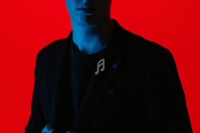 Un homme en costume sombre, éclairé par une lumière bleue sur fond rouge vif.