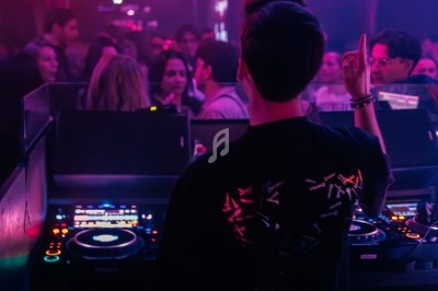 Un DJ mixe dans une boîte de nuit éclairée par des lumières roses, avec une foule dansante en arrière-plan.