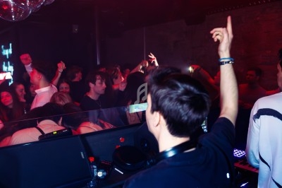 Un DJ mixe sur une scène éclairée par des néons rouges, face à une foule dansante dans une ambiance festive.