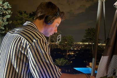Un DJ mixe de la musique en extérieur la nuit, avec une vue éclairée sur une ville en arrière-plan.