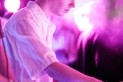 Un DJ en chemise blanche mixe de la musique sur une table de mixage dans une ambiance lumineuse et colorée.