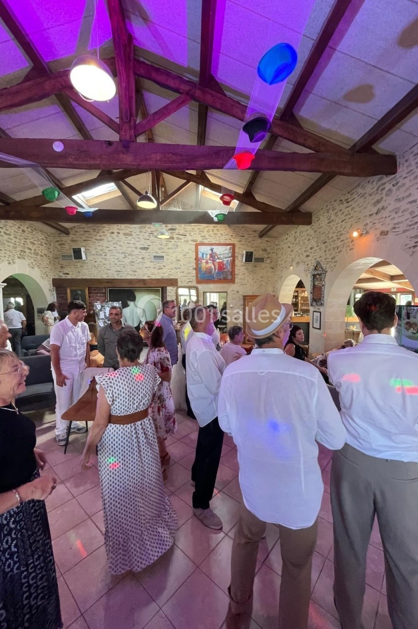 Des personnes dansent et discutent dans une salle avec des murs en pierre, un plafond en poutres apparentes et des lumières…