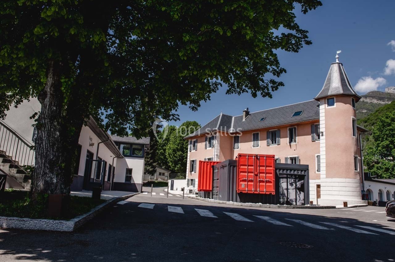 Bâtiment ancien rénové avec une tour, entouré d'arbres, et conteneurs rouges modernes sur le devant.