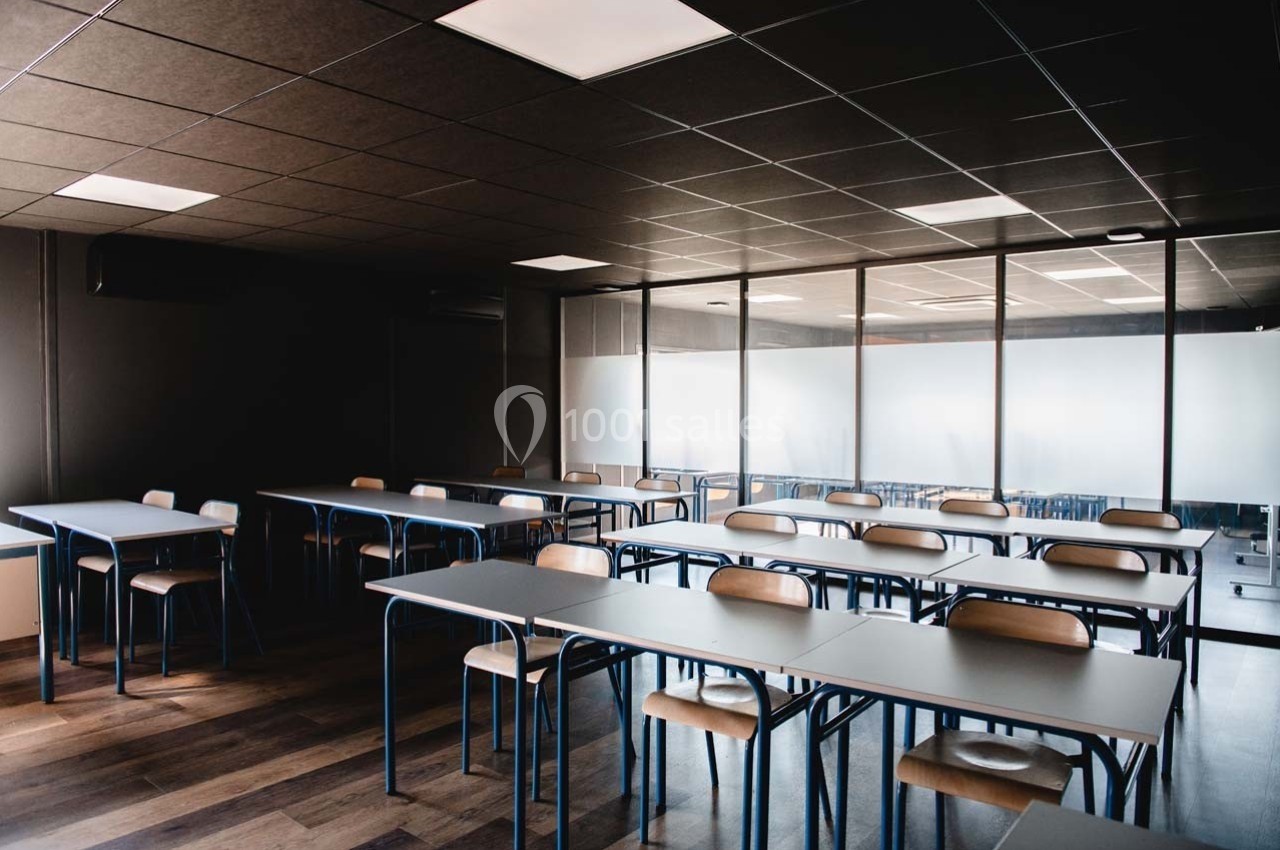 Salle de classe vide avec des tables et des chaises alignées, éclairée par des plafonniers et une lumière naturelle.